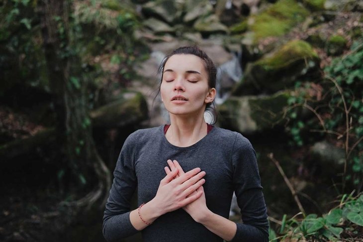 Young woman practicing breathing yoga pranayama outdoors in moss forest on background of waterfall. Unity with nature concept