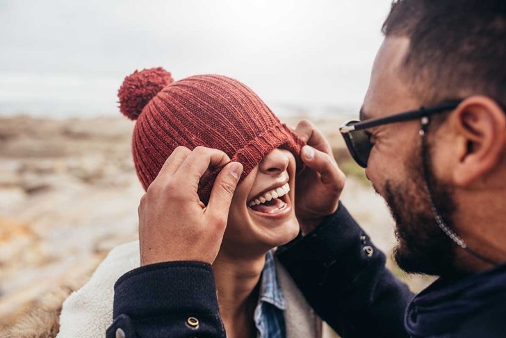 Loving couple having fun outdoors. Man covering eyes of woman with cap.