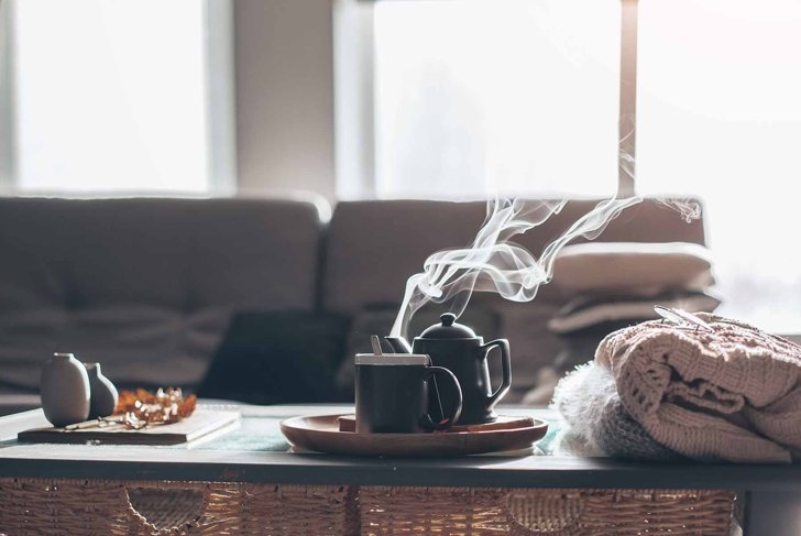 Still life details in home interior of living room. Sweaters and cup of tea with steam on a serving tray on a coffee table. Breakfast over sofa in morning sunlight. Cozy autumn or winter concept.