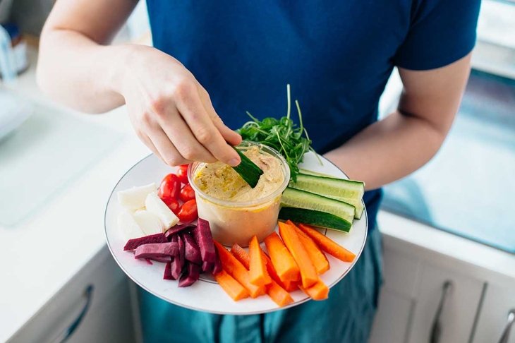 Top view close up man's hand dipping cucumber stick in hummus on the kitchen. Hummus served with raw vegetables on the plate. Healthy food lunch. Vegetarian and vegan food diet. Soft selective focus