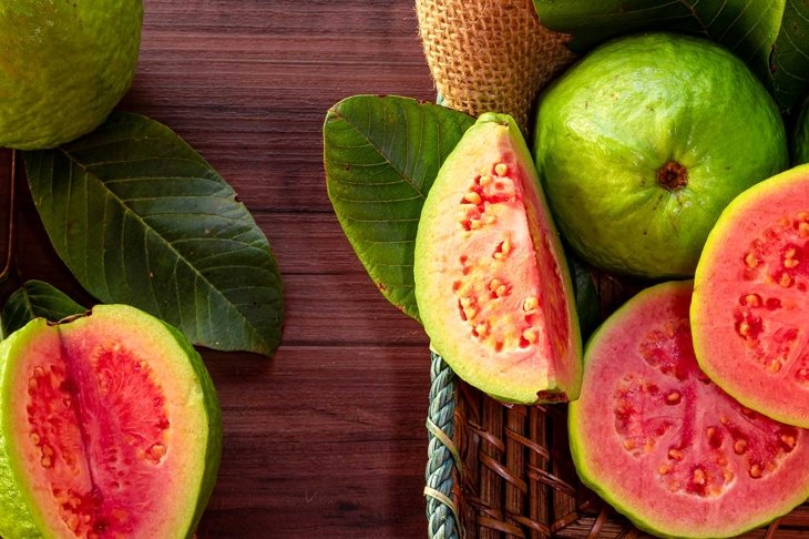 Closeup of a red guava cut in half, in the background several guavas and green leaf