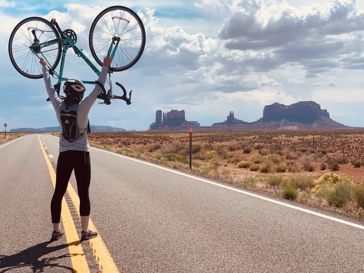 A cyclist standing on an open road holding the bike overhead. 