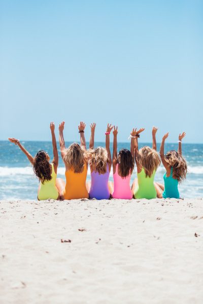 6 women sat on a beach wearing colourful swimsuits.